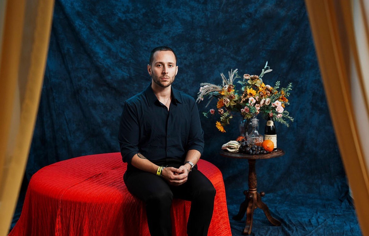 Portrait of Nic Fernandez-Kiray seated on a red-draped table in front of a deep blue backdrop, with a still life of flowers, fruit, and wine beside him